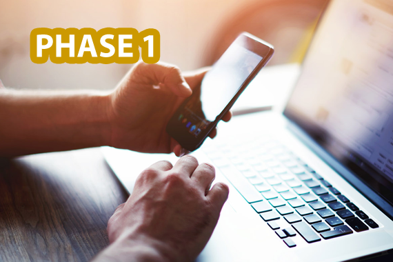 Anmeldestart zum TMS Phase 1 Ansicht von zwei Händen einer Person auf einem Tisch. Die eine Hand hält ein Handy, die andere bewegt den Cursor auf einem Laptop. Oben links ist der Schriftzug 'Phase 1' eingeblendet.