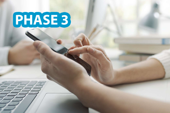Anmeldestart zum TMS Phase 3 Ansicht von zwei Händen einer Person auf einem Tisch mit einem Laptop. Die eine Hand hält ein Handy, die andere bewegt einen Finger auf dem Handybildschirm. Oben links ist der Schriftzug 'Phase 3' eingeblendet.