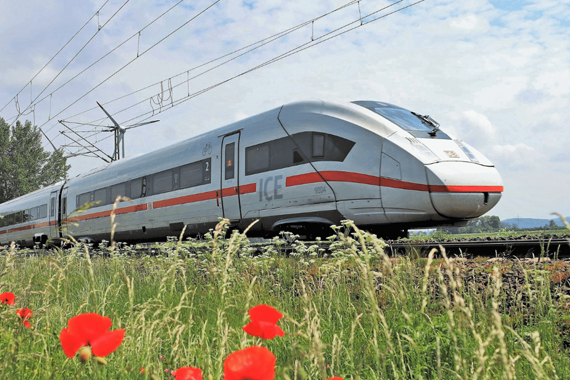Mit der Bahn zum TMS Weißer ICE-Zug mit rotem Streifen auf der Seite, der durch eine Landschaft von links nach rechts fährt. Im Vordergrund sind grüne Gräser und rote Blumen zusehen. Der Himmel ist blau mit weißen Wolken und es sind die Oberleitungen für die Stromversorgung des Zuges zusehen.
