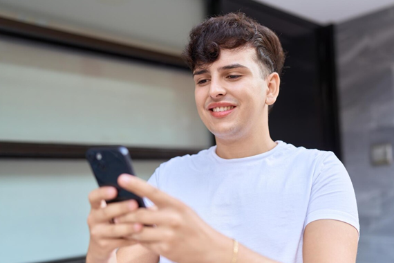 Einladungen für den TMS verfügbar Männliche weiße Person mit dunklen kurzen Haaren und weißen T-Shirt hält mit beiden Händen ein Handy und betrachtet den Inhalt. Im Hintergrund ist eine Hauswand zusehen.