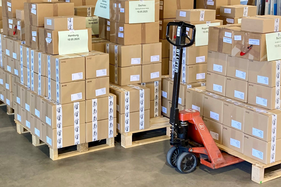 Testheftkartons vom TMS Viele braune Kartons mit Testheften auf Europaletten und einem Hubwagen.